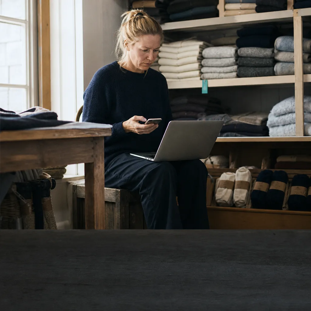 A knitwear founder on a crate in her stockroom, connecting her shop on a laptop